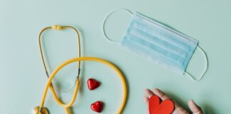 Top view of crop anonymous person hand with red paper heart on table with stethoscope and medical mask for coronavirus prevention