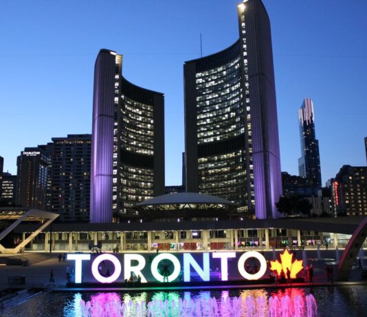 Toronto City Hall illuminated at twilight with colorful Toronto sign in the foreground.
