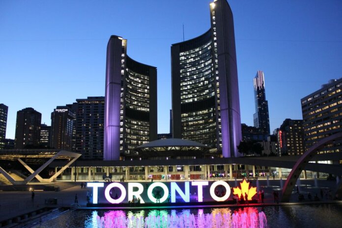 Toronto City Hall illuminated at twilight with colorful Toronto sign in the foreground.