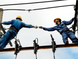Two electrical workers in blue uniforms collaborating on power lines during daytime.