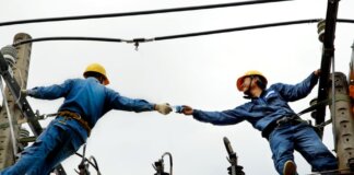 Two electrical workers in blue uniforms collaborating on power lines during daytime.