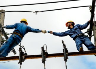 Two electrical workers in blue uniforms collaborating on power lines during daytime.