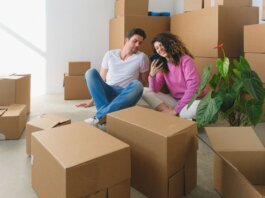 Young couple sitting among cardboard boxes, looking at smartphone, symbolizing moving or relocation.