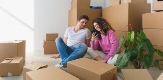 Young couple sitting among cardboard boxes, looking at smartphone, symbolizing moving or relocation.