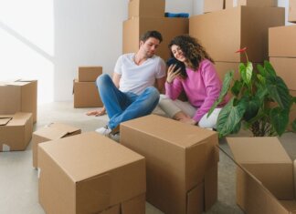 Young couple sitting among cardboard boxes, looking at smartphone, symbolizing moving or relocation.