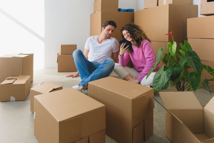 Young couple sitting among cardboard boxes, looking at smartphone, symbolizing moving or relocation.