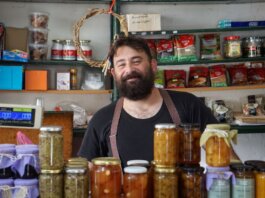A friendly shopkeeper stands behind shelves of homemade preserves.