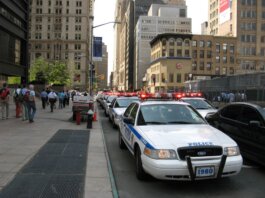 A line of NYPD police cars with flashing lights on a busy New York street, featuring pedestrians and tall buildings.
