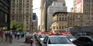 A line of NYPD police cars with flashing lights on a busy New York street, featuring pedestrians and tall buildings.