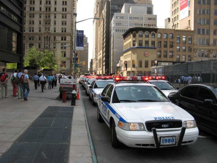 A line of NYPD police cars with flashing lights on a busy New York street, featuring pedestrians and tall buildings. A line of NYPD police cars with flashing lights on a busy New York street, featuring pedestrians and tall buildings.