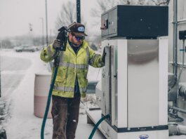 A man wearing a jacket refuels a truck at a gas station during winter with snow falling.