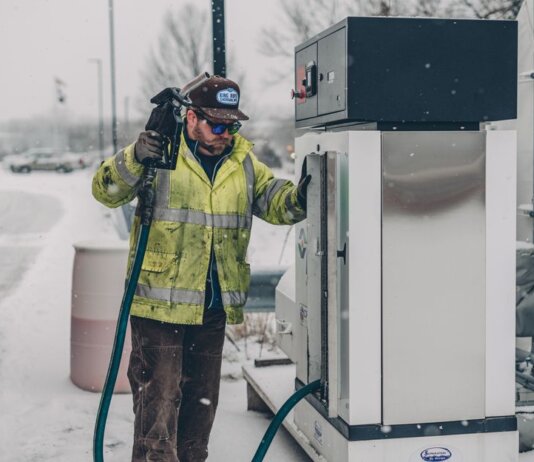 A man wearing a jacket refuels a truck at a gas station during winter with snow falling.