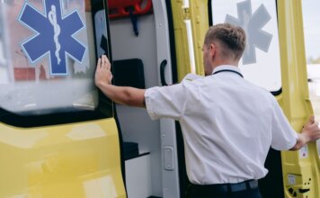 A paramedic in uniform opens the side door of a yellow ambulance during the day.