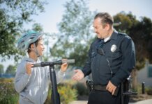 20 лет программе «Молодёжь в полиции» в Дареме A police officer converses with a boy wearing a helmet on a scooter in a sunny outdoor setting.