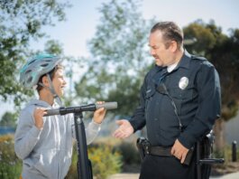 A police officer converses with a boy wearing a helmet on a scooter in a sunny outdoor setting.