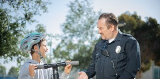 A police officer converses with a boy wearing a helmet on a scooter in a sunny outdoor setting.