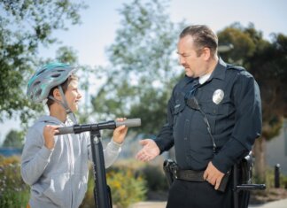 A police officer converses with a boy wearing a helmet on a scooter in a sunny outdoor setting.