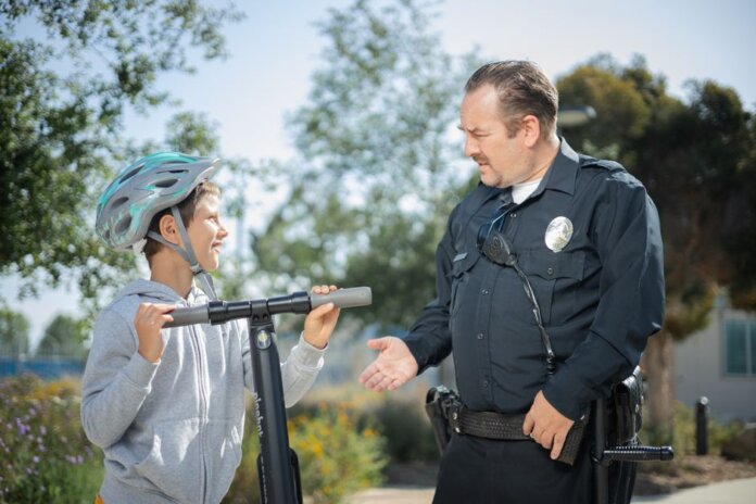 A police officer converses with a boy wearing a helmet on a scooter in a sunny outdoor setting. A police officer converses with a boy wearing a helmet on a scooter in a sunny outdoor setting.
