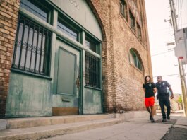 A police officer escorts a handcuffed individual past a brick building.