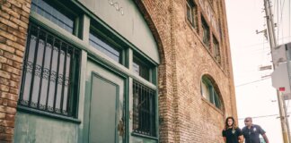 A police officer escorts a handcuffed individual past a brick building.