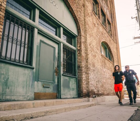 A police officer escorts a handcuffed individual past a brick building.
