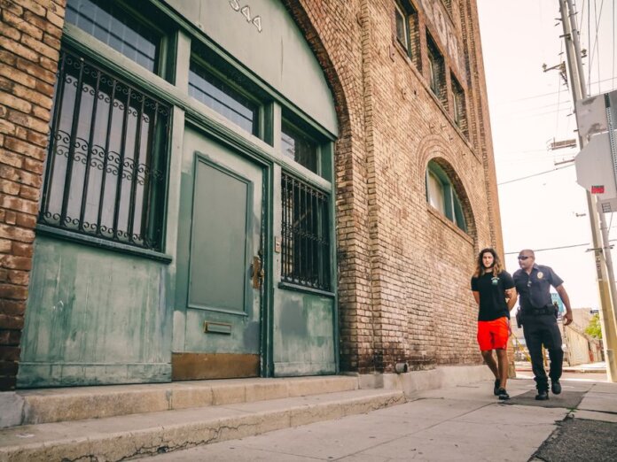 A police officer escorts a handcuffed individual past a brick building. A police officer escorts a handcuffed individual past a brick building.