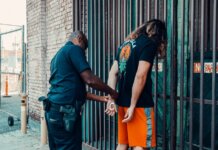 A police officer handcuffing a man against a metal fence in an urban area.