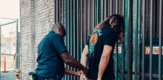 A police officer handcuffing a man against a metal fence in an urban area.