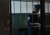 Охранник обвинён в сексуальном насилии во время дежурства A security officer in uniform standing behind bars in a dimly lit room, evoking themes of law enforcement and confinement.