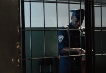 Охранник обвинён в сексуальном насилии во время дежурства A security officer in uniform standing behind bars in a dimly lit room, evoking themes of law enforcement and confinement.