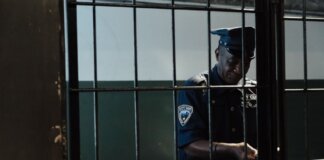 A security officer in uniform standing behind bars in a dimly lit room, evoking themes of law enforcement and confinement.