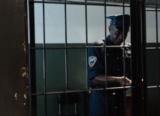 Охранник обвинён в сексуальном насилии во время дежурства A security officer in uniform standing behind bars in a dimly lit room, evoking themes of law enforcement and confinement.
