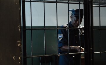 Охранник обвинён в сексуальном насилии во время дежурства A security officer in uniform standing behind bars in a dimly lit room, evoking themes of law enforcement and confinement.
