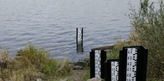A water level measurement post standing in a lake, surrounded by rocks and vegetation.