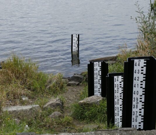 A water level measurement post standing in a lake, surrounded by rocks and vegetation.