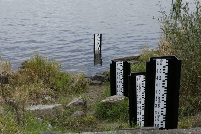A water level measurement post standing in a lake, surrounded by rocks and vegetation.