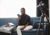 Booking.com: 84% путешественников Азии видят в поездках способ перезагрузки Adult man sits on sofa with open luggage, holding passport, preparing for travel, vlogging.