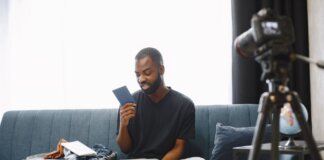 Adult man sits on sofa with open luggage, holding passport, preparing for travel, vlogging.
