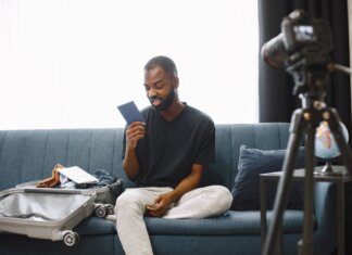 Booking.com: 84% путешественников Азии видят в поездках способ перезагрузки Adult man sits on sofa with open luggage, holding passport, preparing for travel, vlogging.