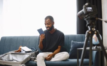 Booking.com: 84% путешественников Азии видят в поездках способ перезагрузки Adult man sits on sofa with open luggage, holding passport, preparing for travel, vlogging.