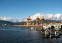 Bright day at a container port with orange cranes and mountain backdrop.