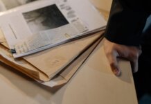 Close-up of a hand resting on a desk near a stack of investigative documents and newspaper clippings.
