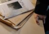 Close-up of a hand resting on a desk near a stack of investigative documents and newspaper clippings.