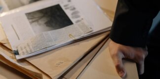 Close-up of a hand resting on a desk near a stack of investigative documents and newspaper clippings.