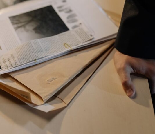 Close-up of a hand resting on a desk near a stack of investigative documents and newspaper clippings.