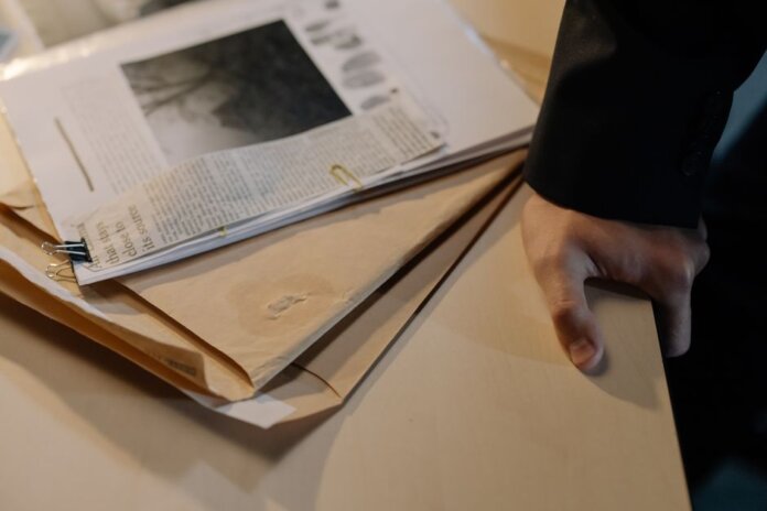 Close-up of a hand resting on a desk near a stack of investigative documents and newspaper clippings. Close-up of a hand resting on a desk near a stack of investigative documents and newspaper clippings.