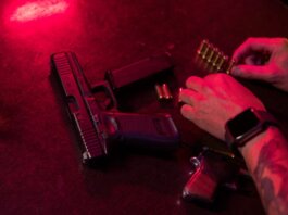 Dramatic close-up of a tattooed hand loading bullets beside a handgun and whiskey glass.