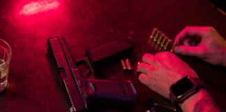 Dramatic close-up of a tattooed hand loading bullets beside a handgun and whiskey glass.