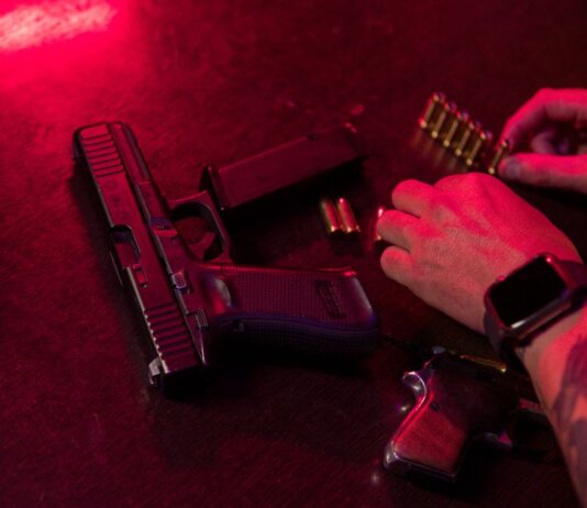 Dramatic close-up of a tattooed hand loading bullets beside a handgun and whiskey glass.