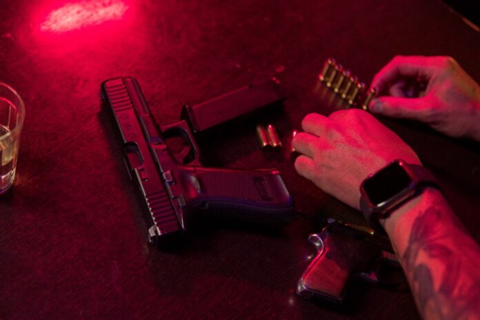 Dramatic close-up of a tattooed hand loading bullets beside a handgun and whiskey glass. Dramatic close-up of a tattooed hand loading bullets beside a handgun and whiskey glass.